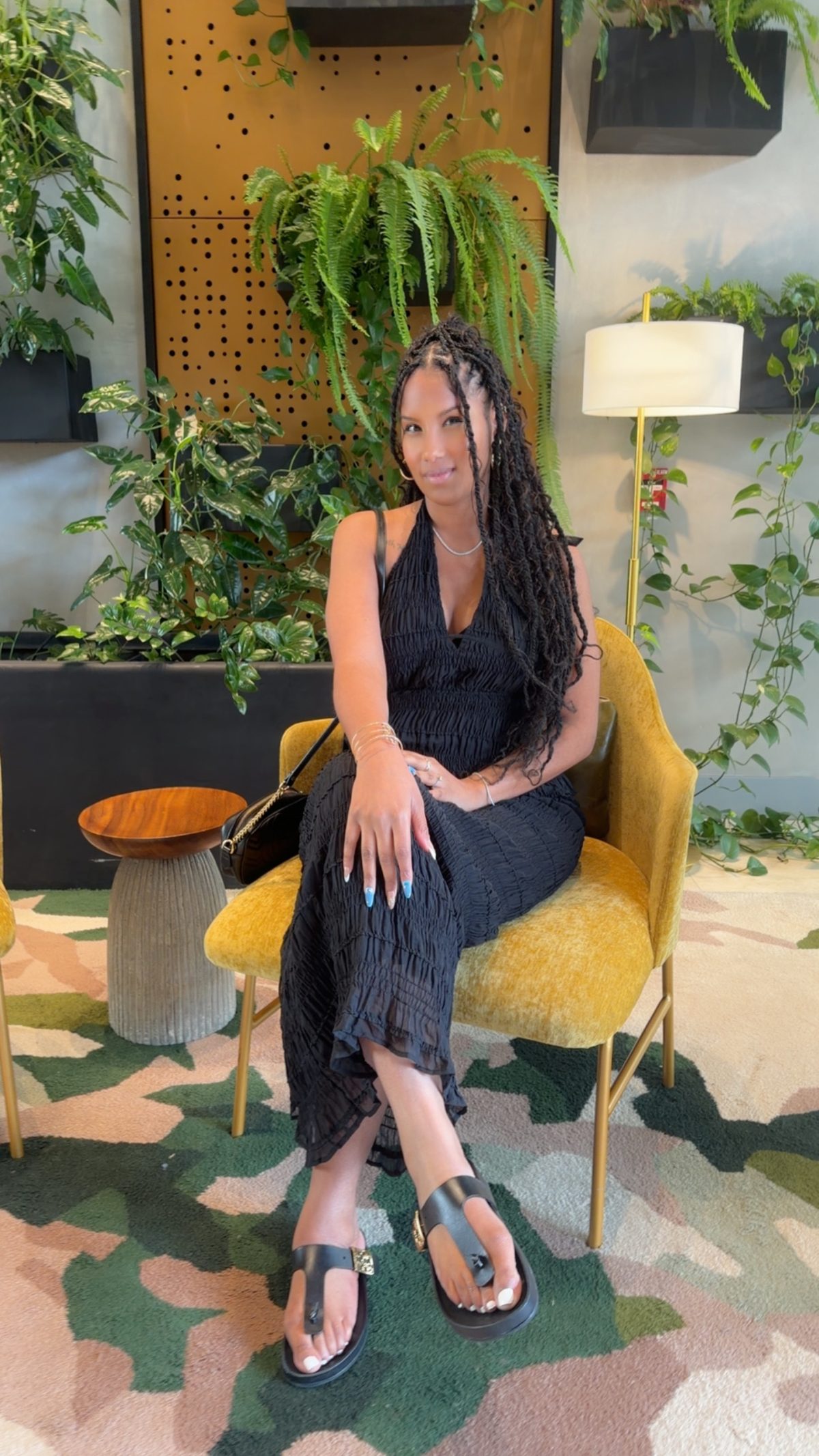 Juelle, smiling, seated in a mustard velvet chair in a black halter dress, with ferns and greenery behind her.
