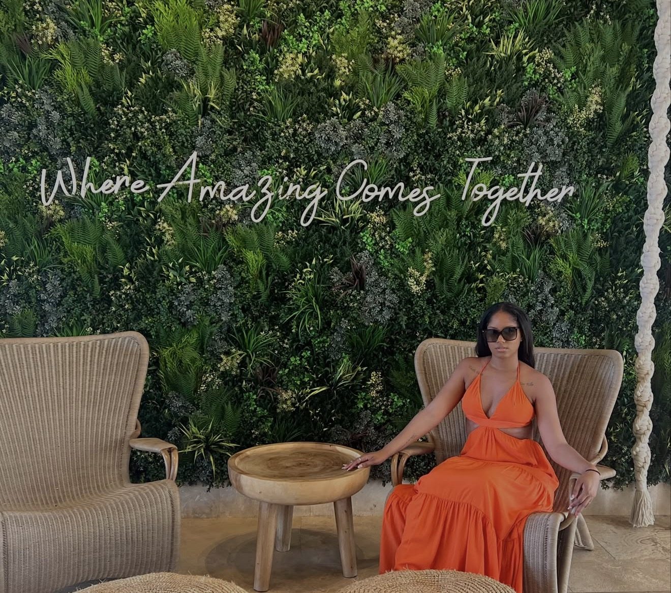 Juelle in an orange maxi dress and sunglasses, seated in a rattan chair in front of a lush green living wall at a resort.